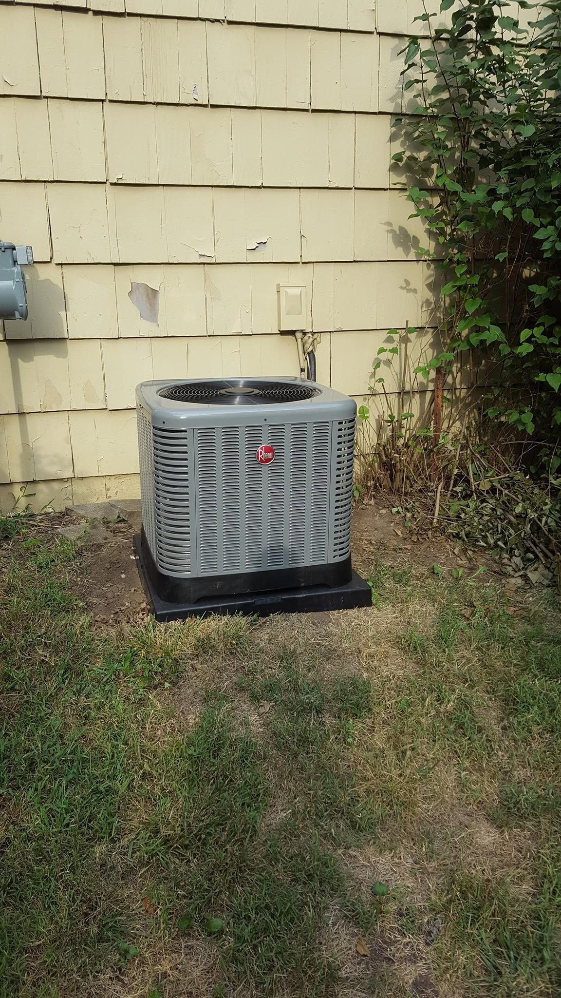 Outdoor air conditioner unit beside a beige house wall and grass, with bushes on the right