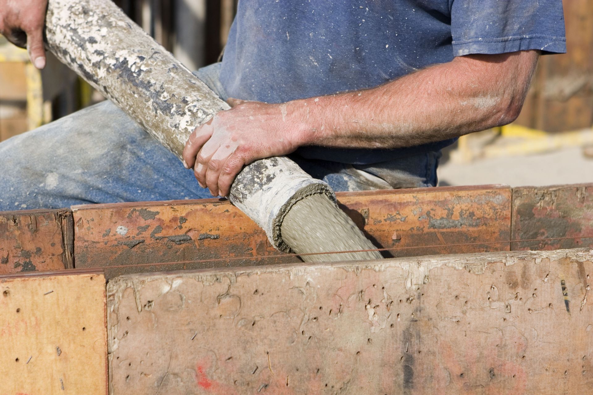 A worker in a blue shirt directs a concrete pump hose into a wooden frame at a construction site.