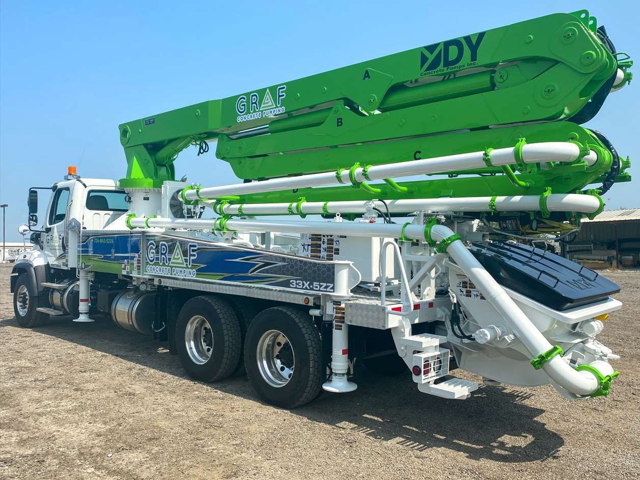 A bright green and white DY concrete boom pump truck parked on a dirt lot under a clear blue sky.