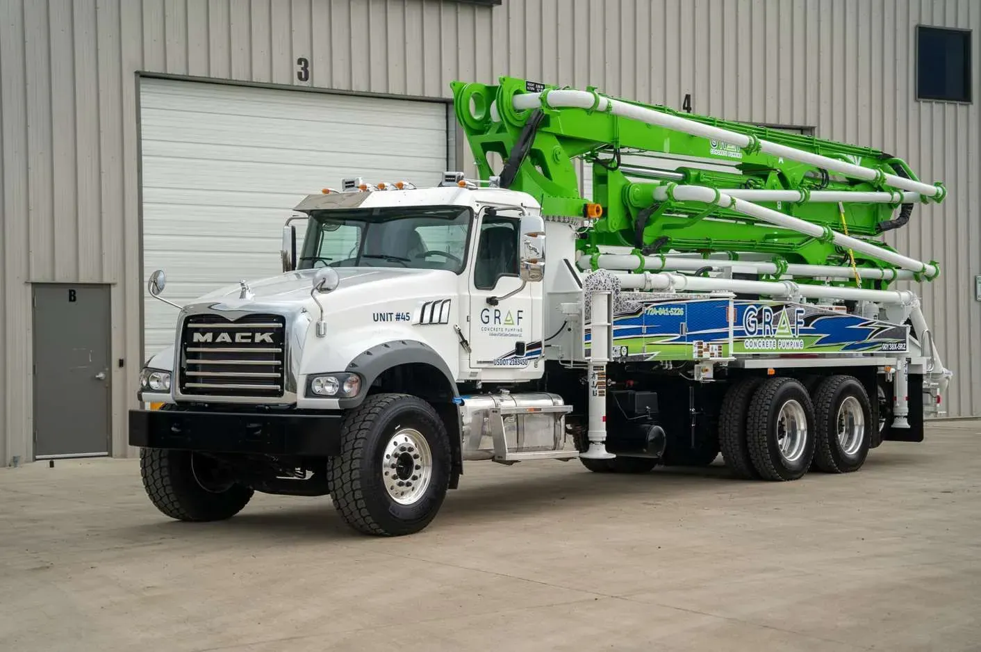 A white Mack truck with a large, lime green concrete pump boom mounted on the back, parked in front of a warehouse.