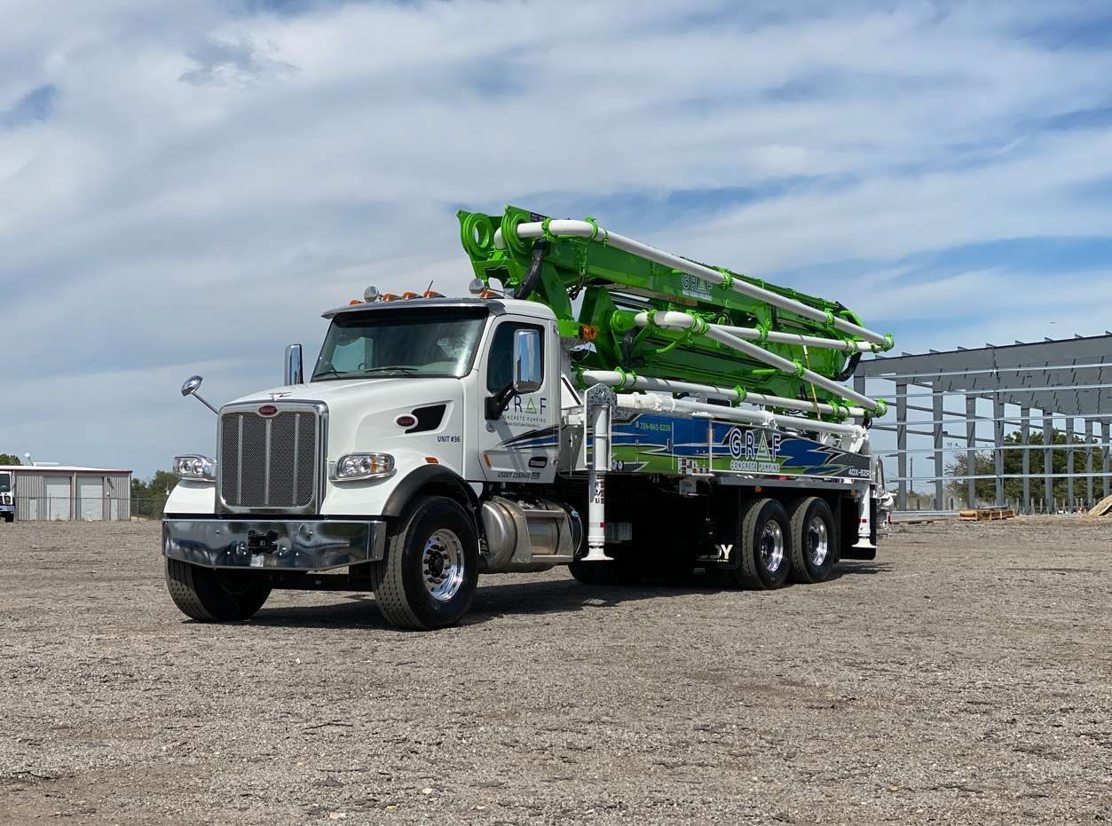 A white industrial concrete pump truck with a folded green boom arm parked on a gravel construction site.