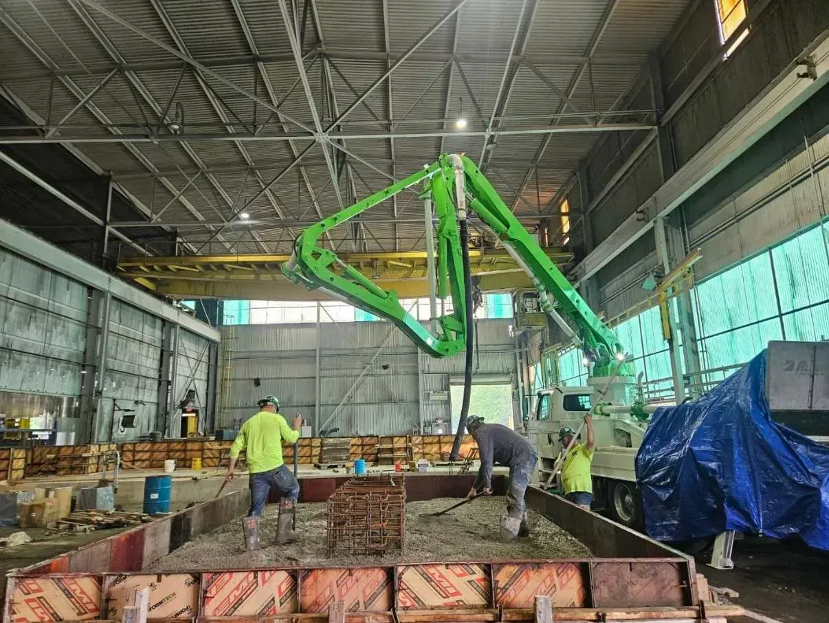 Workers in high-visibility vests operate a green concrete pump truck inside a large, industrial construction site.
