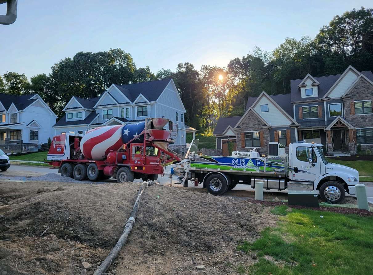 A red, white, and blue cement truck pours concrete into a pump truck in a suburban neighborhood during sunset.