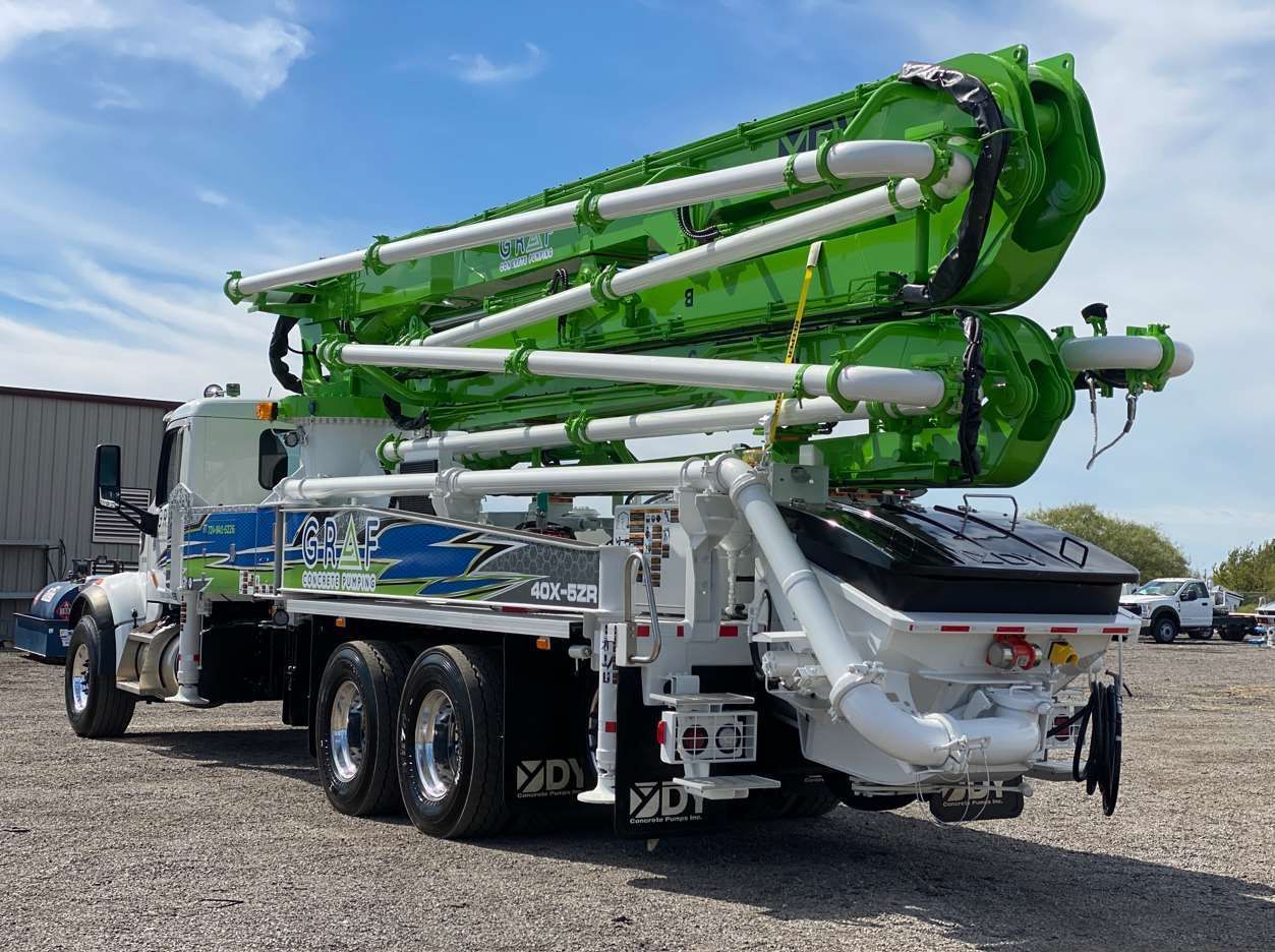 A white and lime-green concrete pump truck parked on a gravel lot under a blue, cloudy sky.