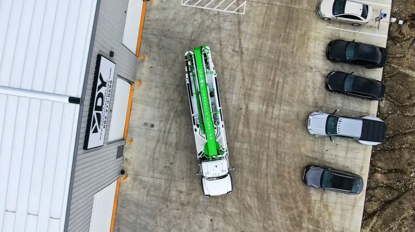 An aerial view shows a white and green truck parked next to a building, with several cars parked in a row nearby.