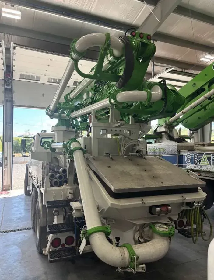 A white concrete pump truck with a folded green articulated boom arm parked inside a large garage.