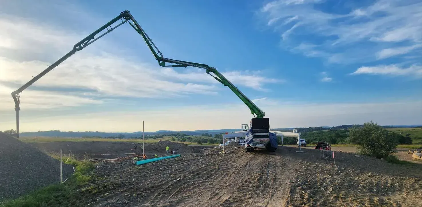 A long-armed green concrete pump truck extends its boom over a construction site under a partly cloudy blue sky.