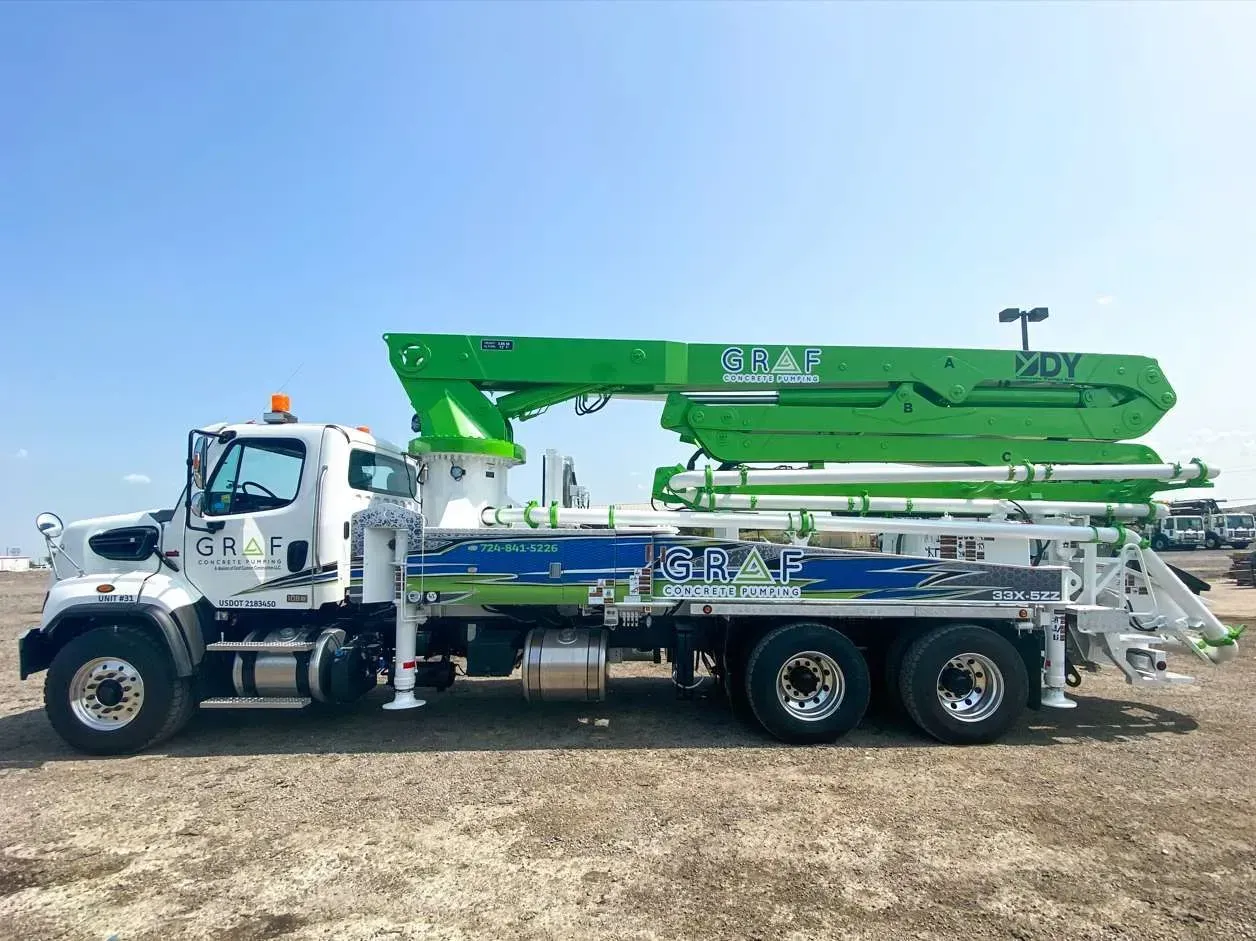 A white concrete pump truck with a long, folded lime green boom arm parked on a gravel lot under a clear blue sky.