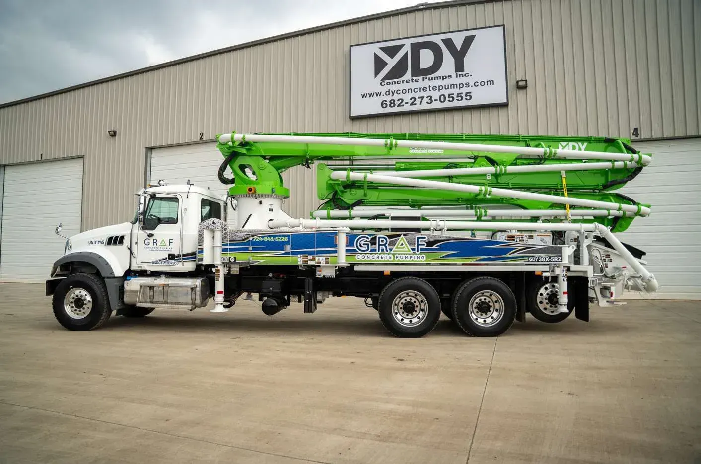 A white concrete pump truck with a folded lime green boom arm parked in front of a DY Concrete Pumps building.