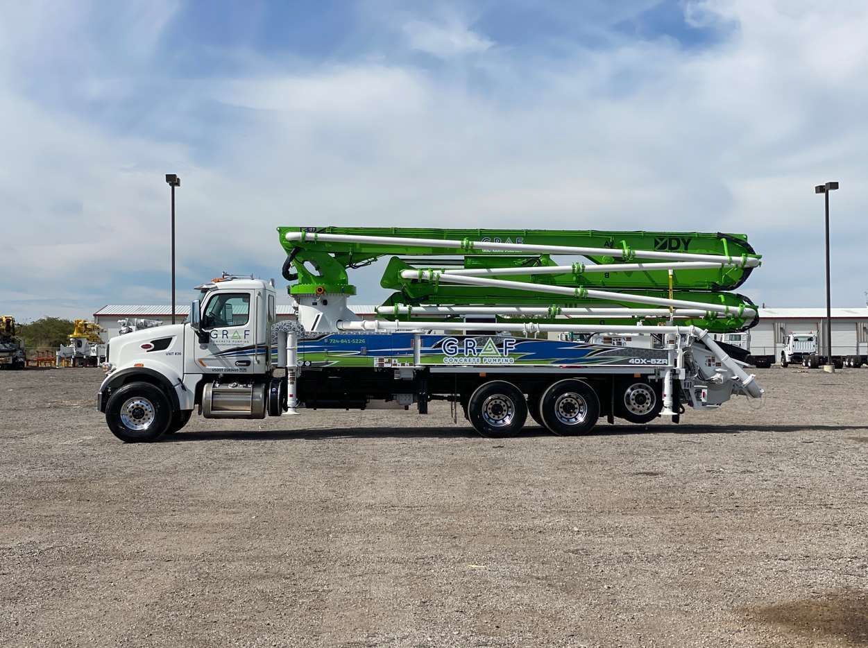 A white Peterbilt truck with a folded, bright green concrete pump boom mounted on the back in a gravel lot.