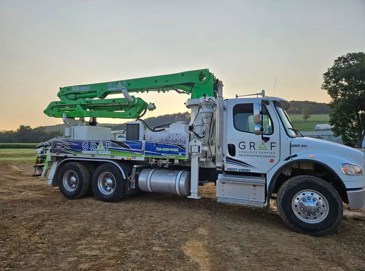 A white and green concrete pump truck parked on a dirt lot at dusk, with rolling hills in the background.