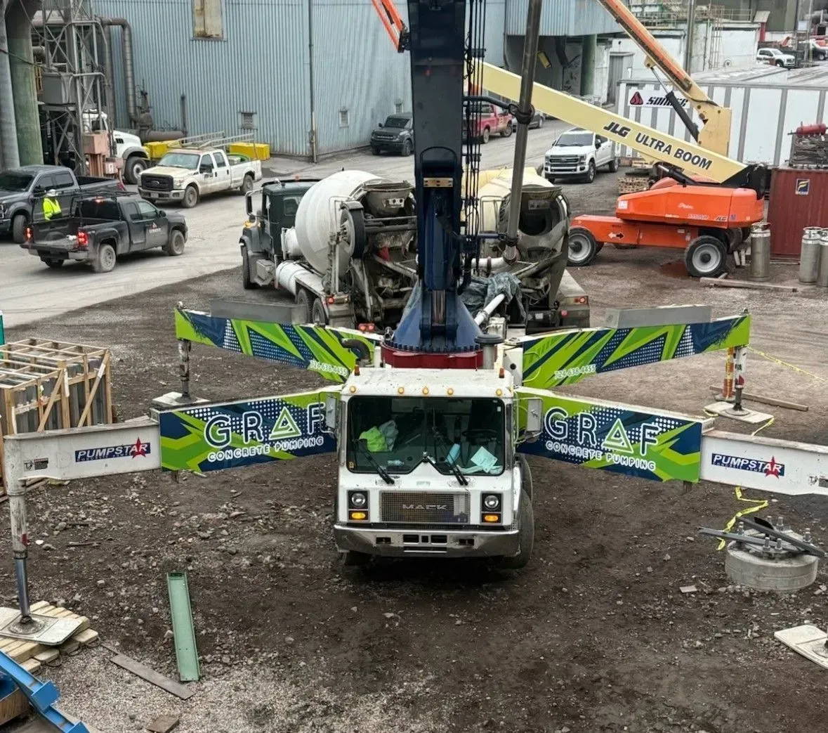 A concrete pump truck with extended outriggers positioned in a construction yard with other vehicles and machinery.