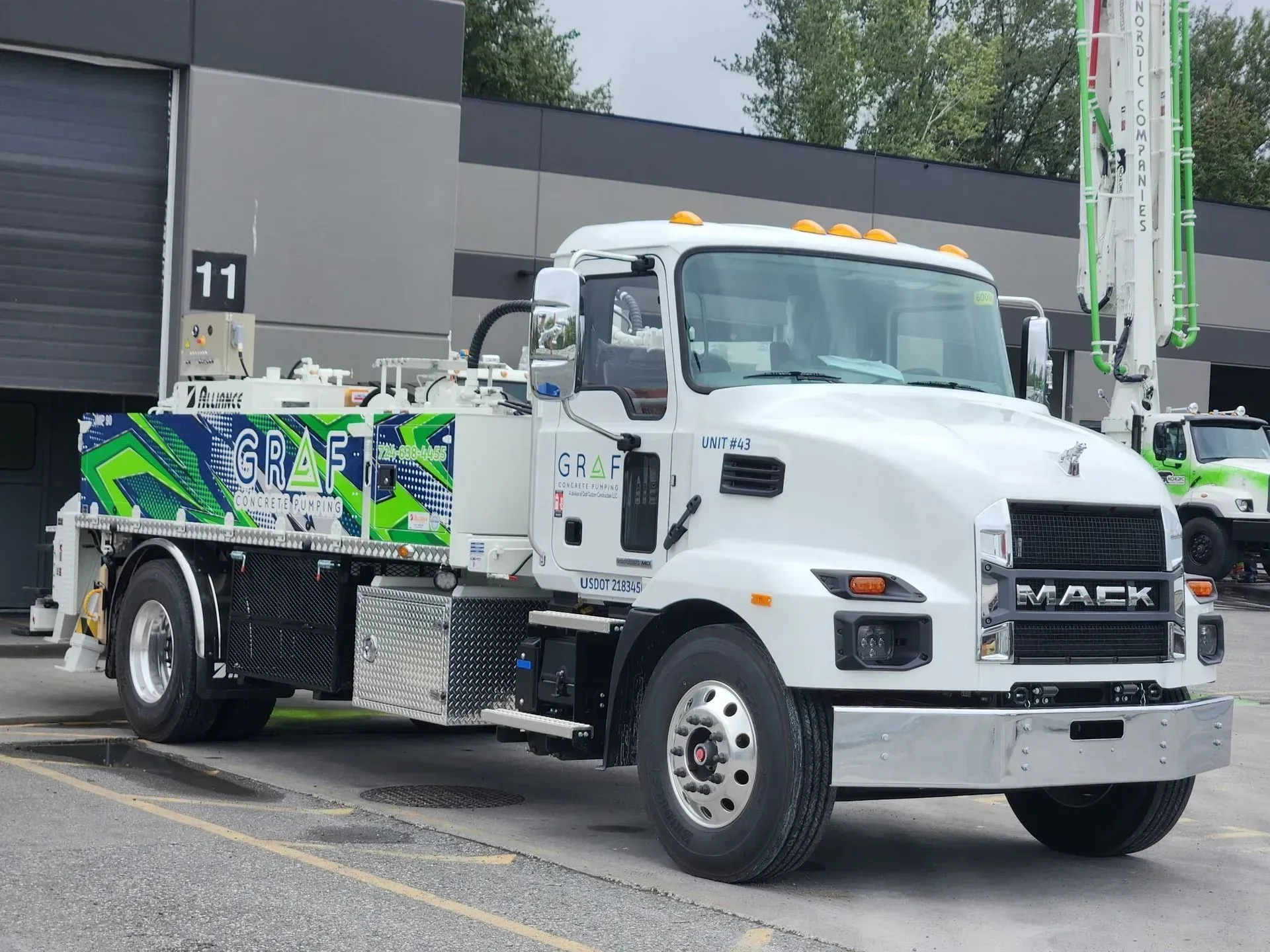 A white Mack concrete pump truck parked in front of a commercial building labeled with the number 11.