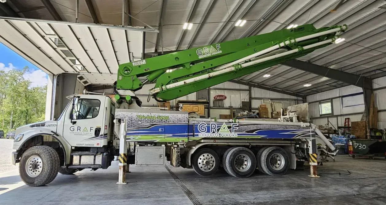 A green and white concrete pump truck parked inside a bright workshop with its boom arm partially extended.