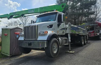A white Peterbilt concrete pump truck with a green boom extended outdoors, parked next to a portable toilet.