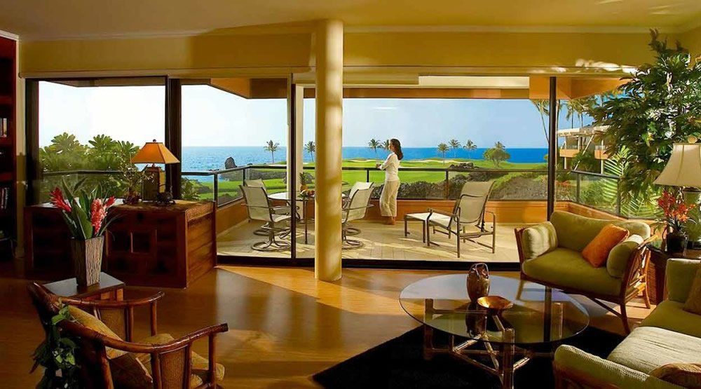 A living room with a woman standing on a balcony overlooking the ocean
