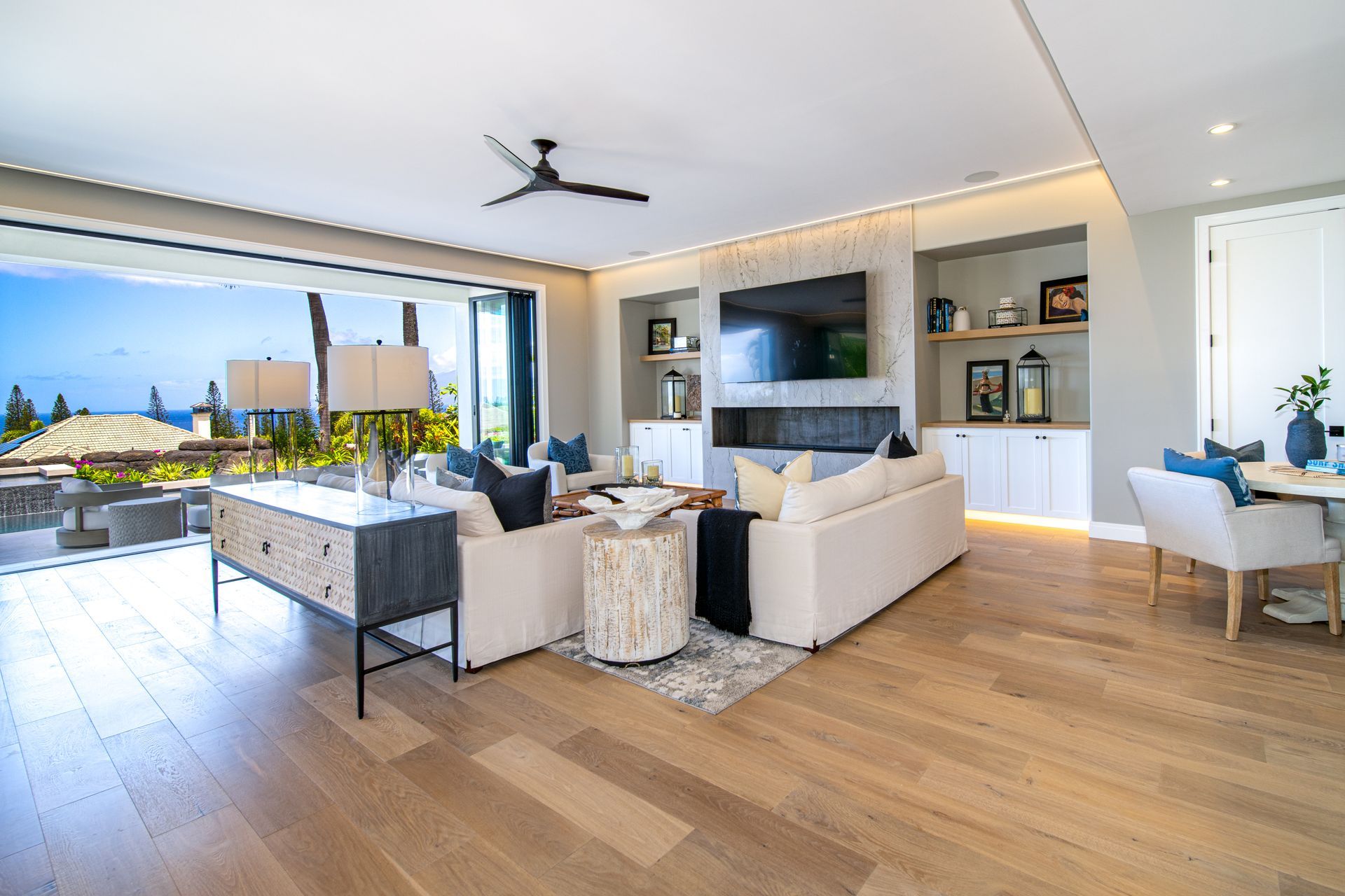 A living room with hardwood floors , white furniture , a flat screen tv and a ceiling fan.