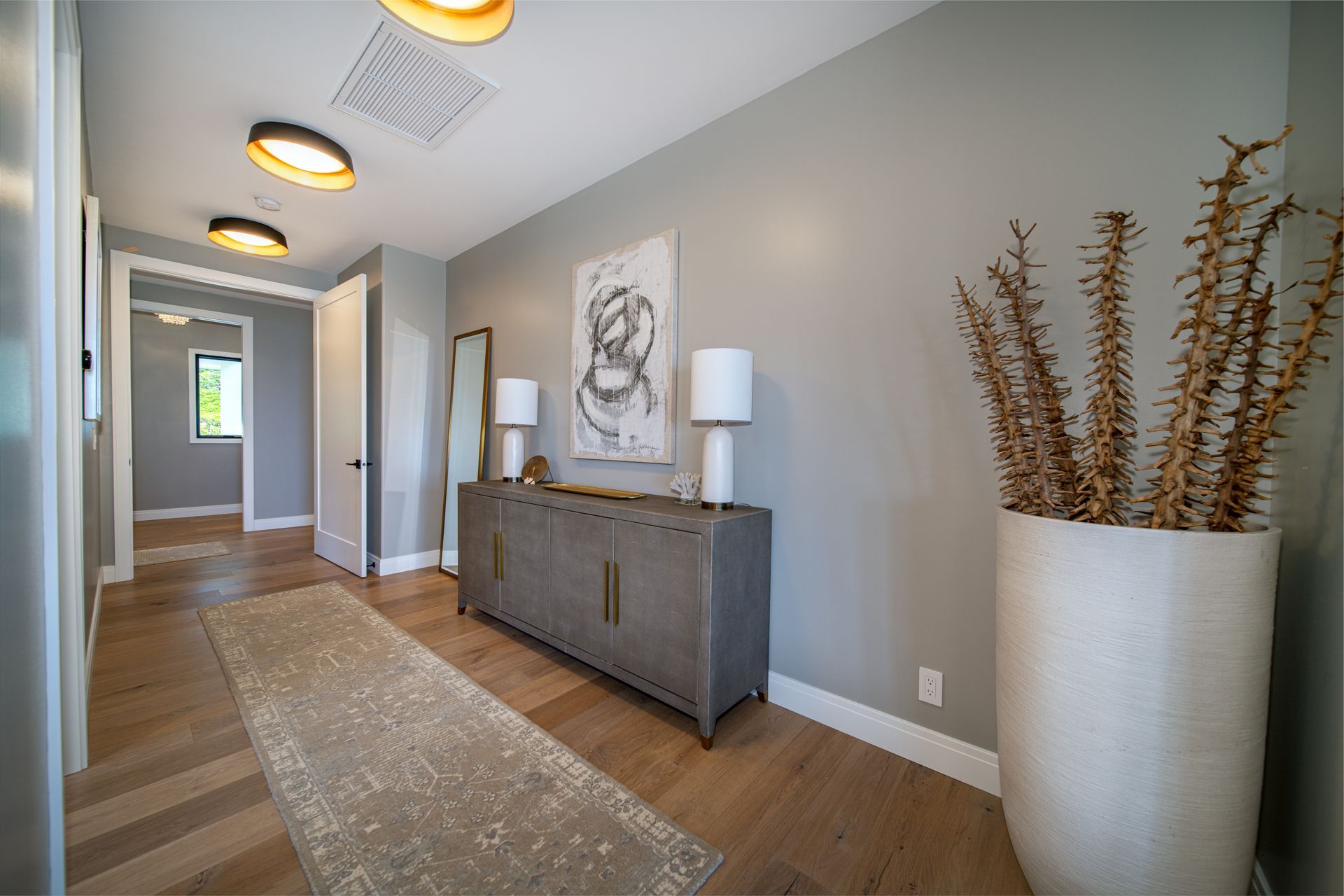 A hallway with a dresser , lamps , a vase and a painting on the wall.