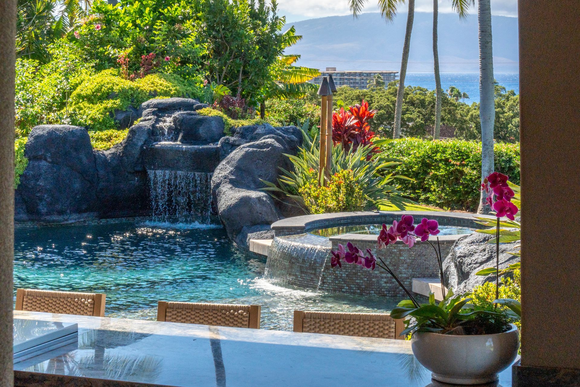 A swimming pool with a waterfall in the background and a table and chairs in the foreground.
