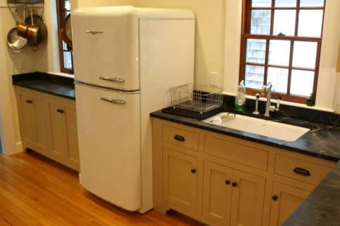 Cream-colored vintage refrigerator in a kitchen with tan cabinets, a sink, and a window.