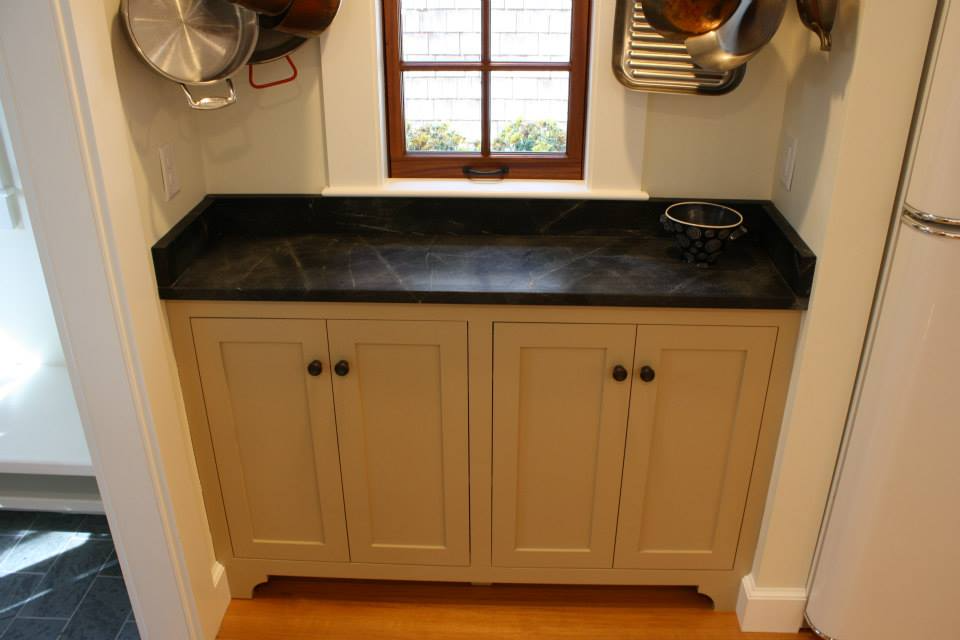 Cream-colored kitchen cabinet with black countertop under a small window, pots hanging above.