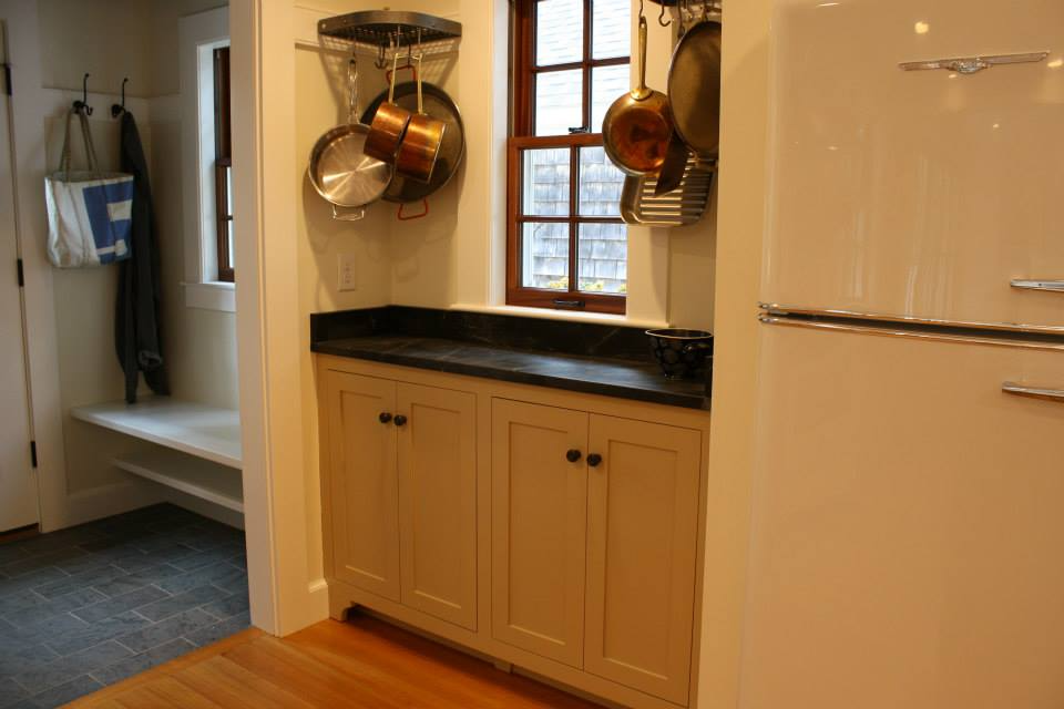 Cream-colored kitchen cabinets with dark countertop, copper pots hanging on wall, small window, and white refrigerator.