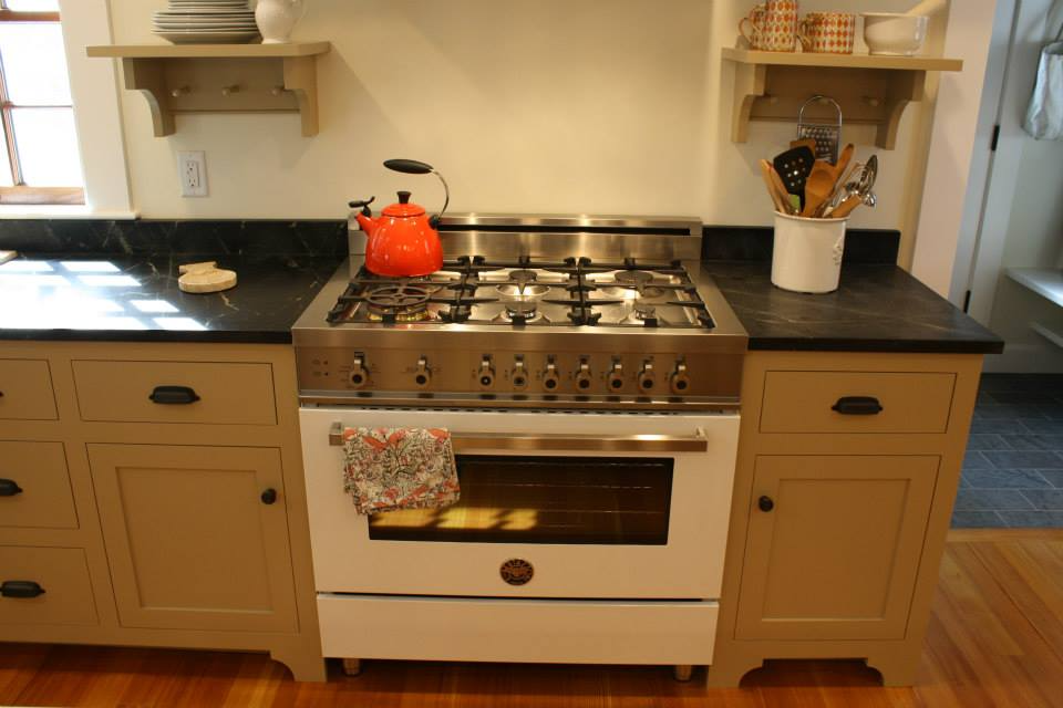 Kitchen with a white oven, stainless steel stovetop, tan cabinets, and black countertops. An orange kettle sits on top.