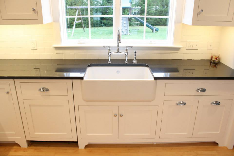 White farmhouse kitchen with a large sink, black countertop, and a window overlooking a yard.