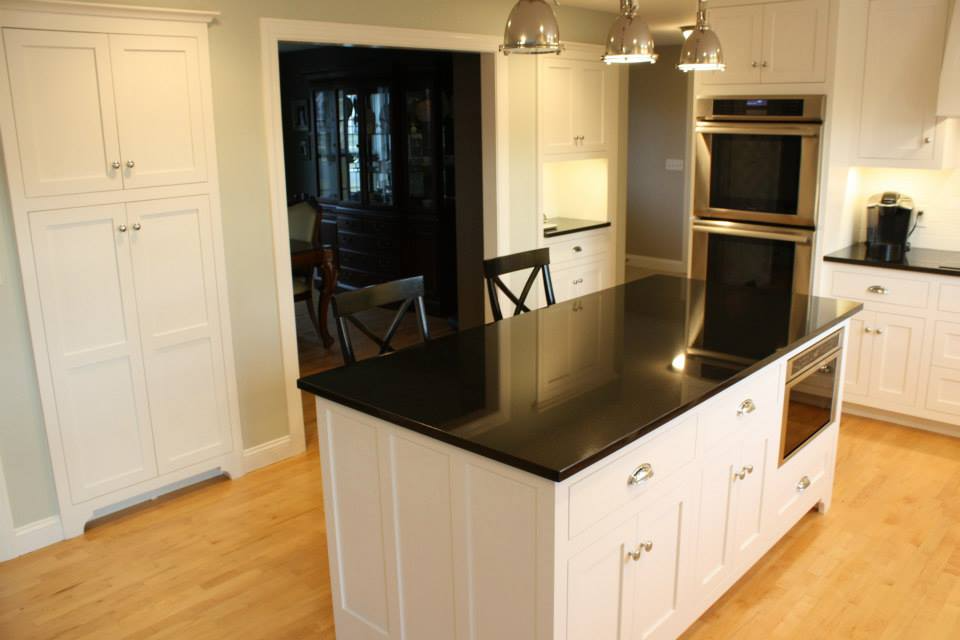 White kitchen with black countertop island and built-in appliances.  Blonde wood floor,  light fixtures, open doorway.