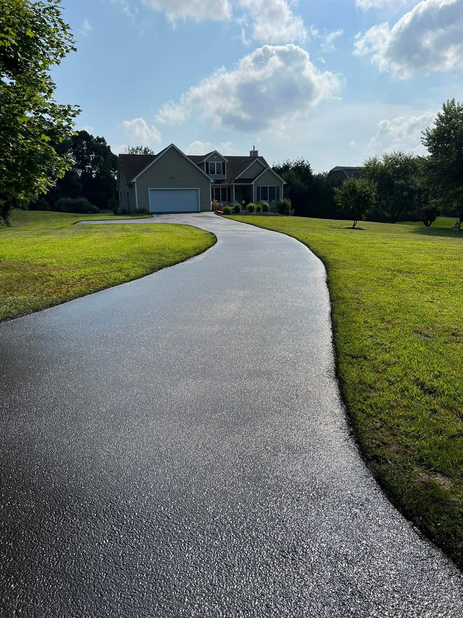 A long, freshly paved asphalt driveway leads up to a residential house under a bright, partly cloudy sky.