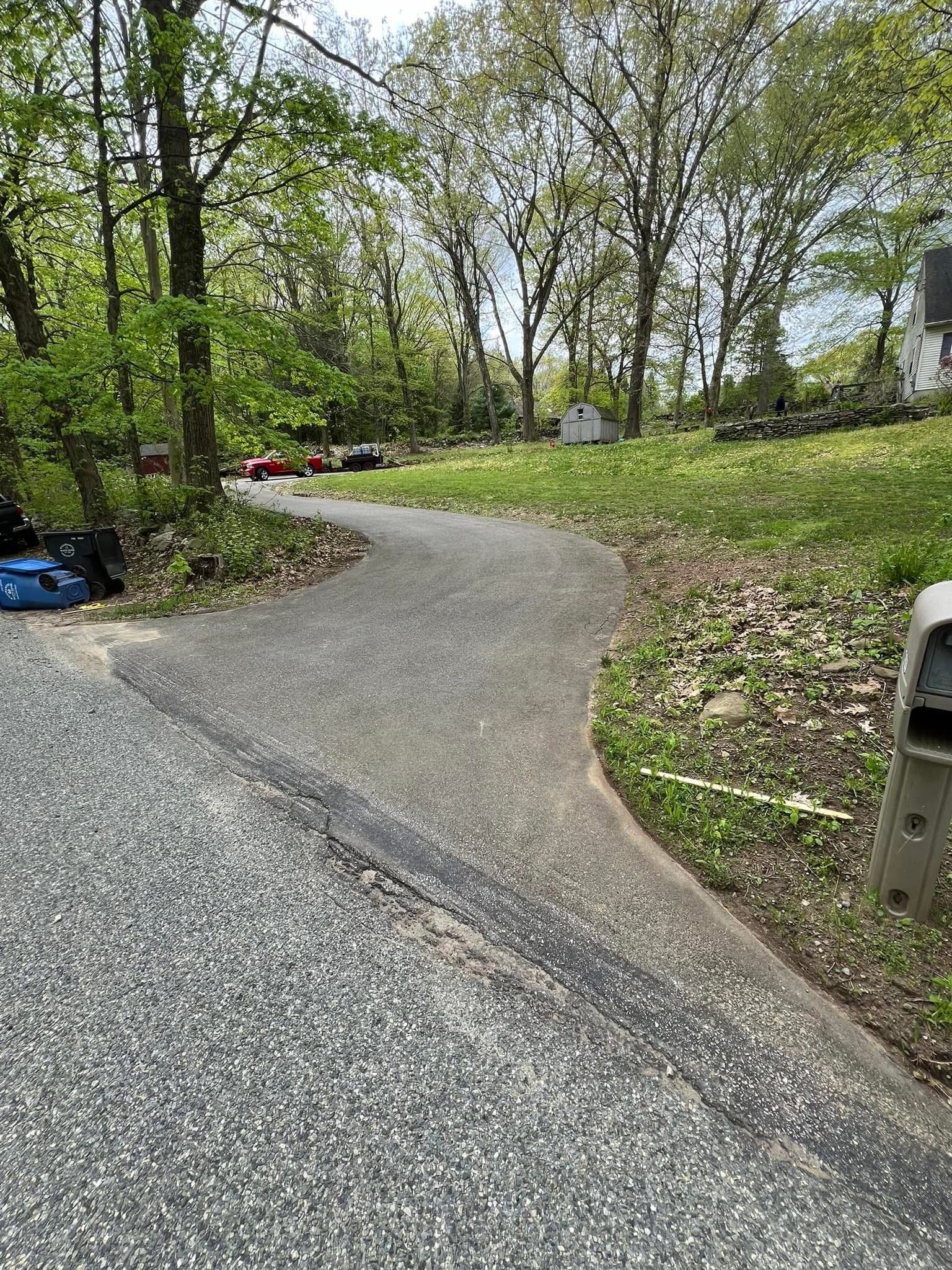 A paved, curved driveway leads from a coarse gravel surface toward a grassy yard with trees under a bright sky.
