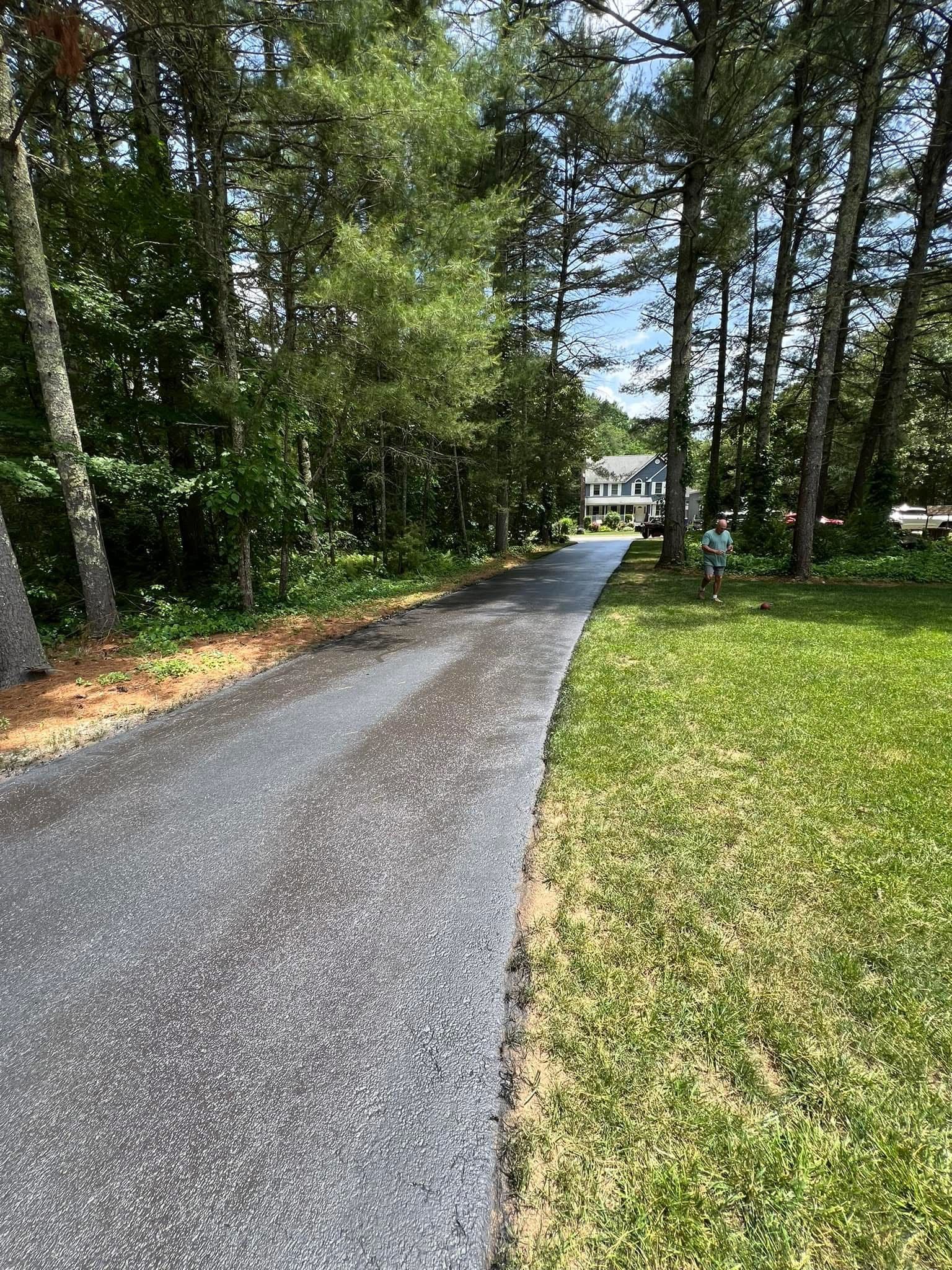 A newly paved asphalt driveway leads through a wooded area toward a house in the distance.