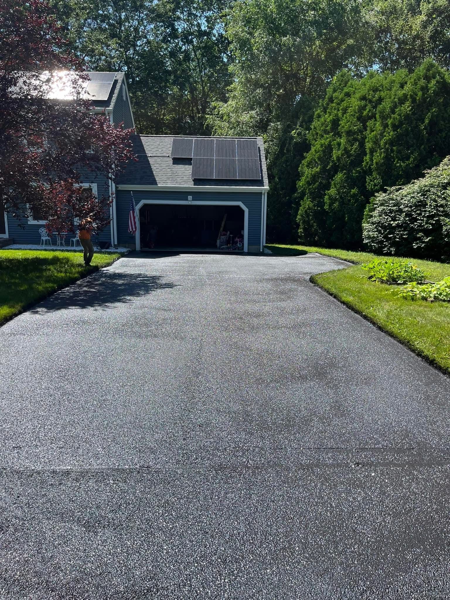 A newly seal-coated dark gray asphalt driveway leads to a suburban garage with rooftop solar panels on a sunny day.