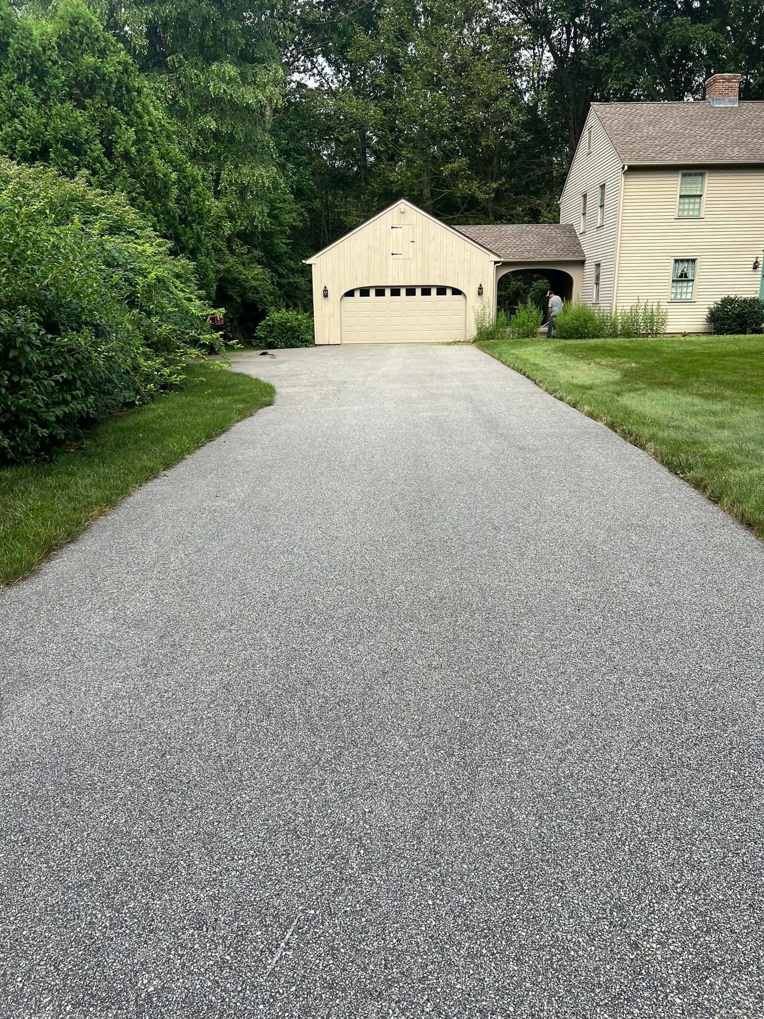 A gravel driveway leading toward a detached garage and the side of a light-colored home surrounded by trees.