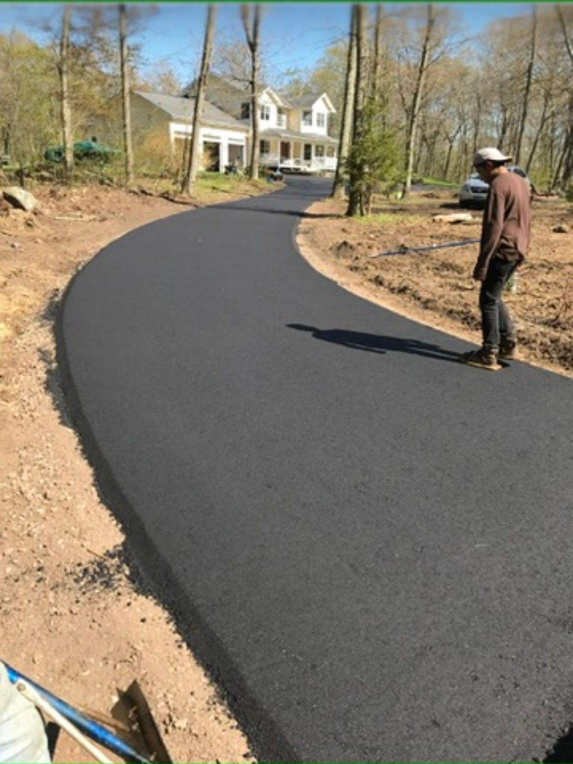 A person stands on a freshly paved, curved asphalt driveway leading to a multi-story house surrounded by trees.