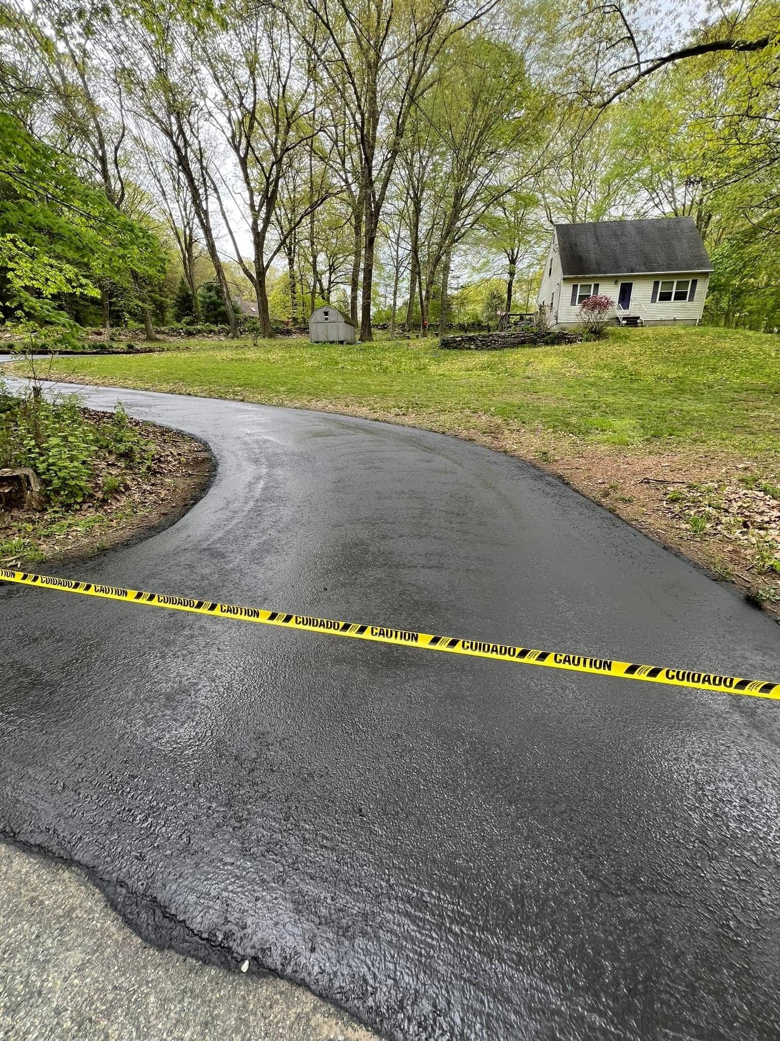 A yellow caution tape stretches across a freshly paved, dark asphalt driveway leading toward a house in a wooded area.