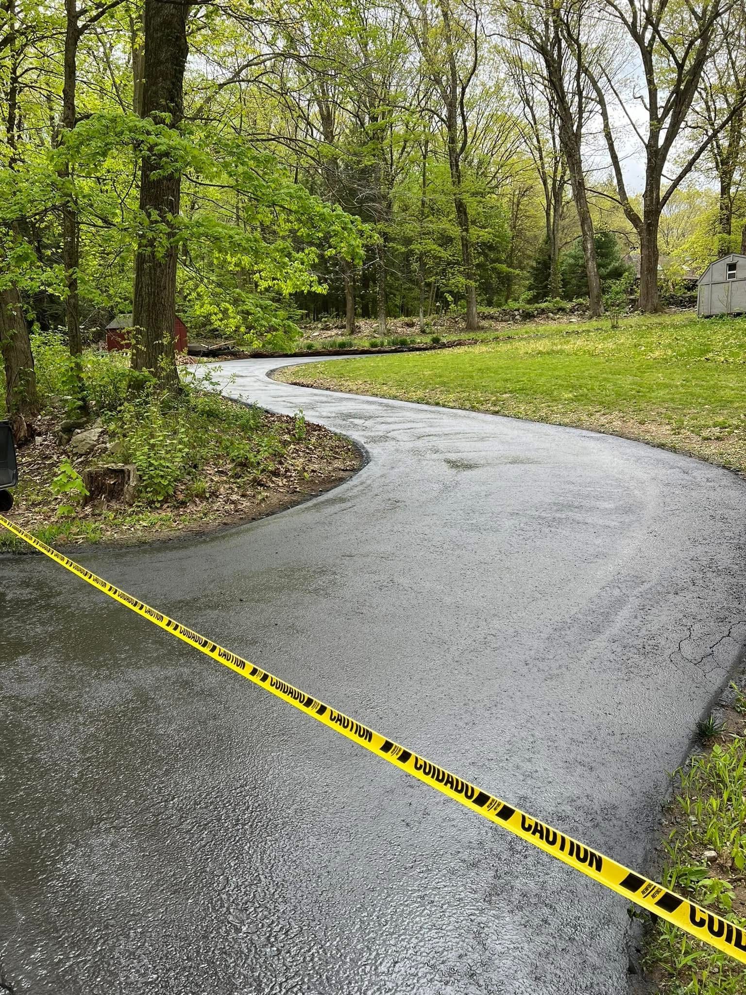 A newly paved, winding driveway bordered by trees and grass, blocked off by a strip of yellow caution tape.