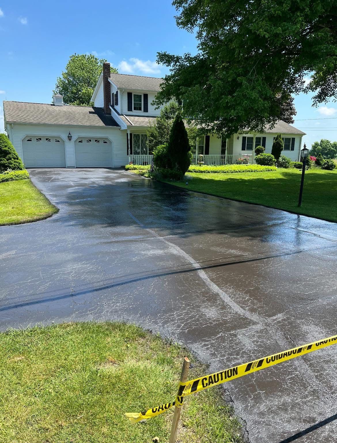 A white house with a two-car garage and a recently sealed, shiny black asphalt driveway blocked by yellow caution tape.