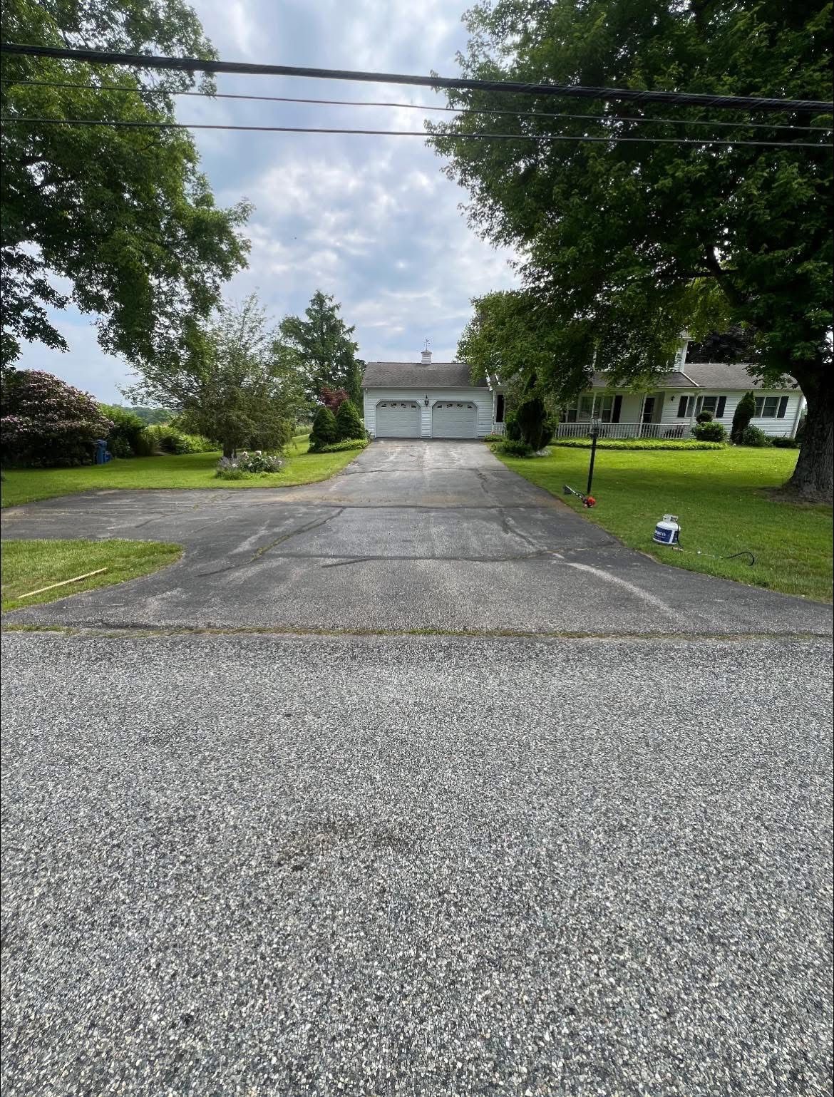 A gravel driveway leads to an asphalt path heading toward a white house and a detached two-car garage under a cloudy sky.