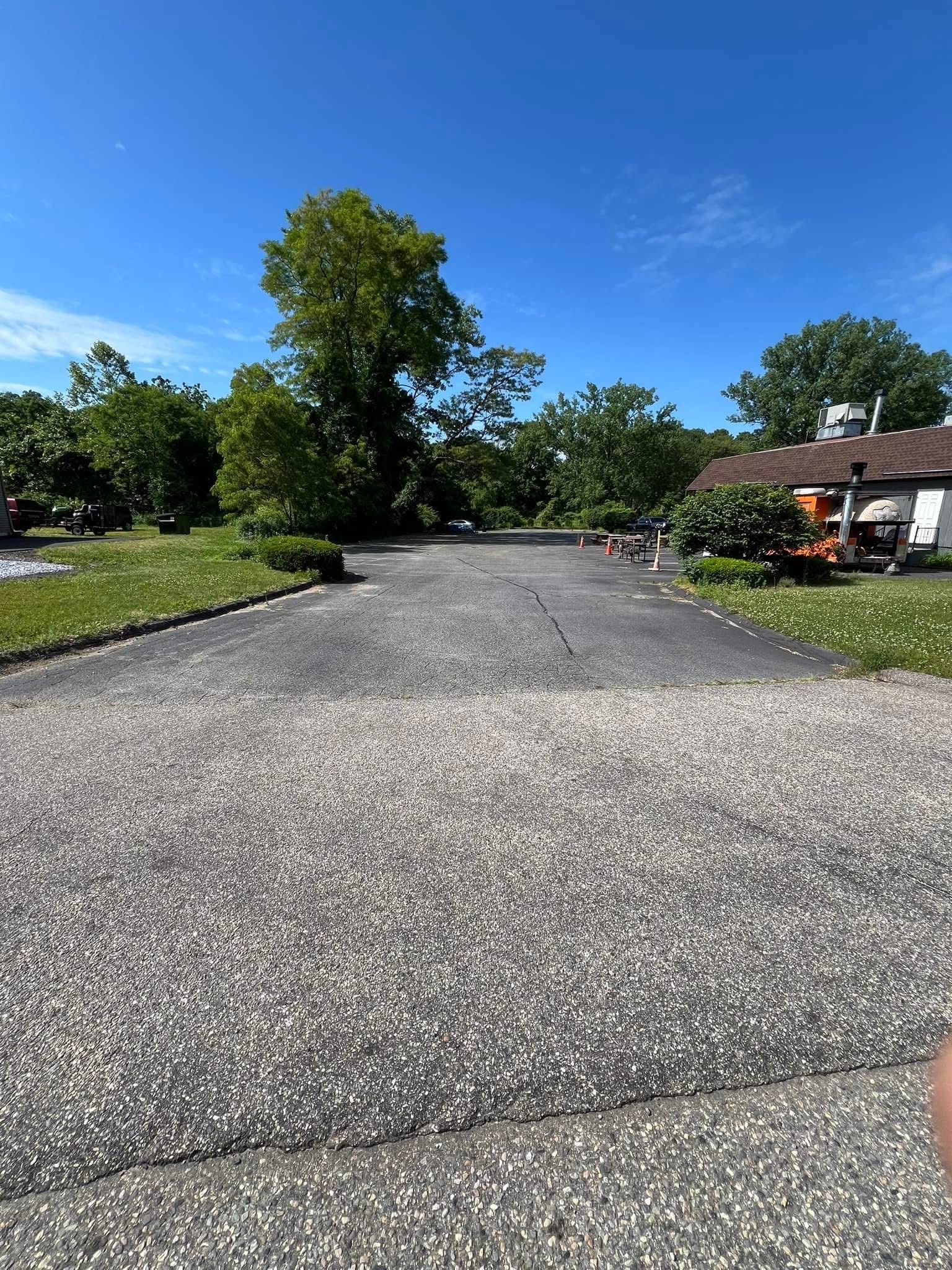 A paved driveway leads to a parking area surrounded by trees and a building under a bright blue sky.