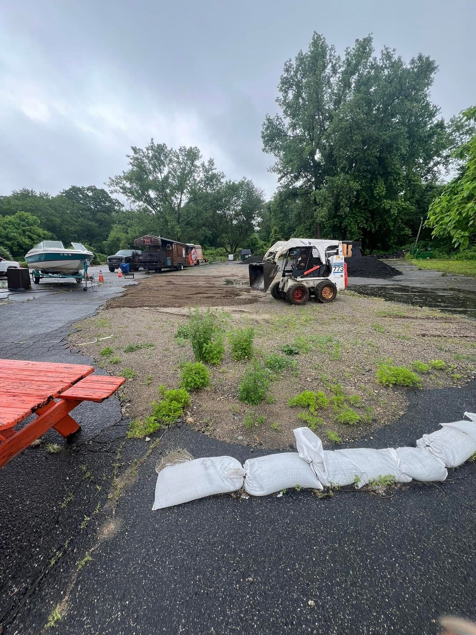 A white skid steer loader operates on a gravel lot near a parked boat and red picnic table under a cloudy sky.