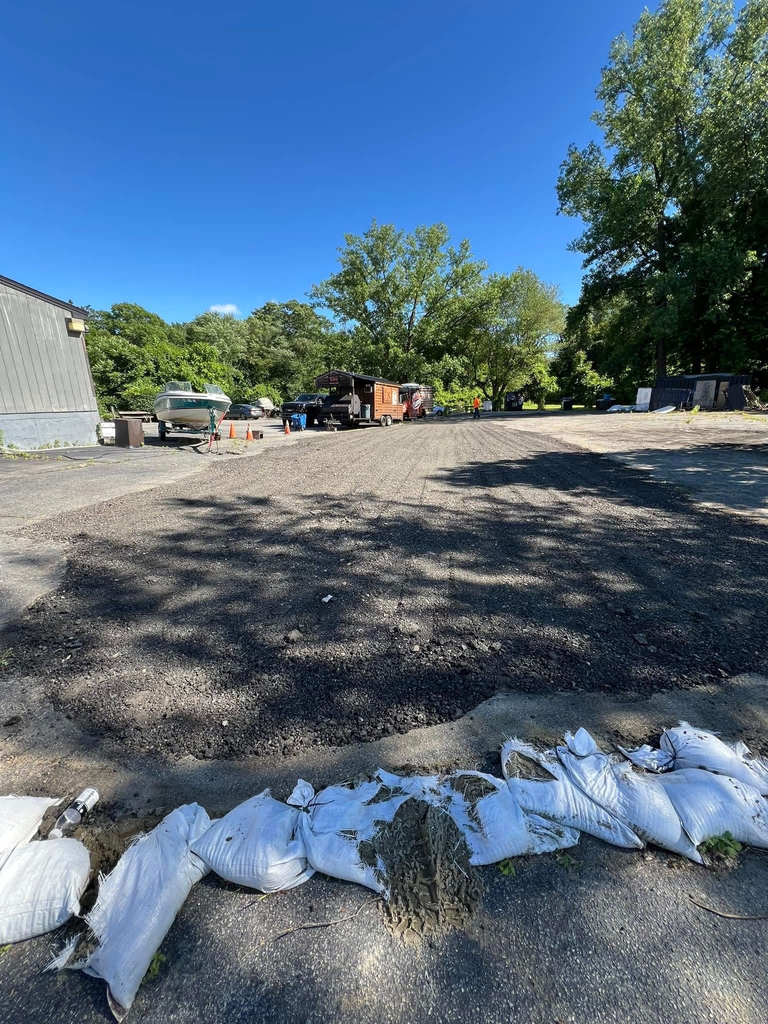 A gravel-covered area under a clear blue sky, bordered by white sandbags in the foreground, with trees and vehicles behind.