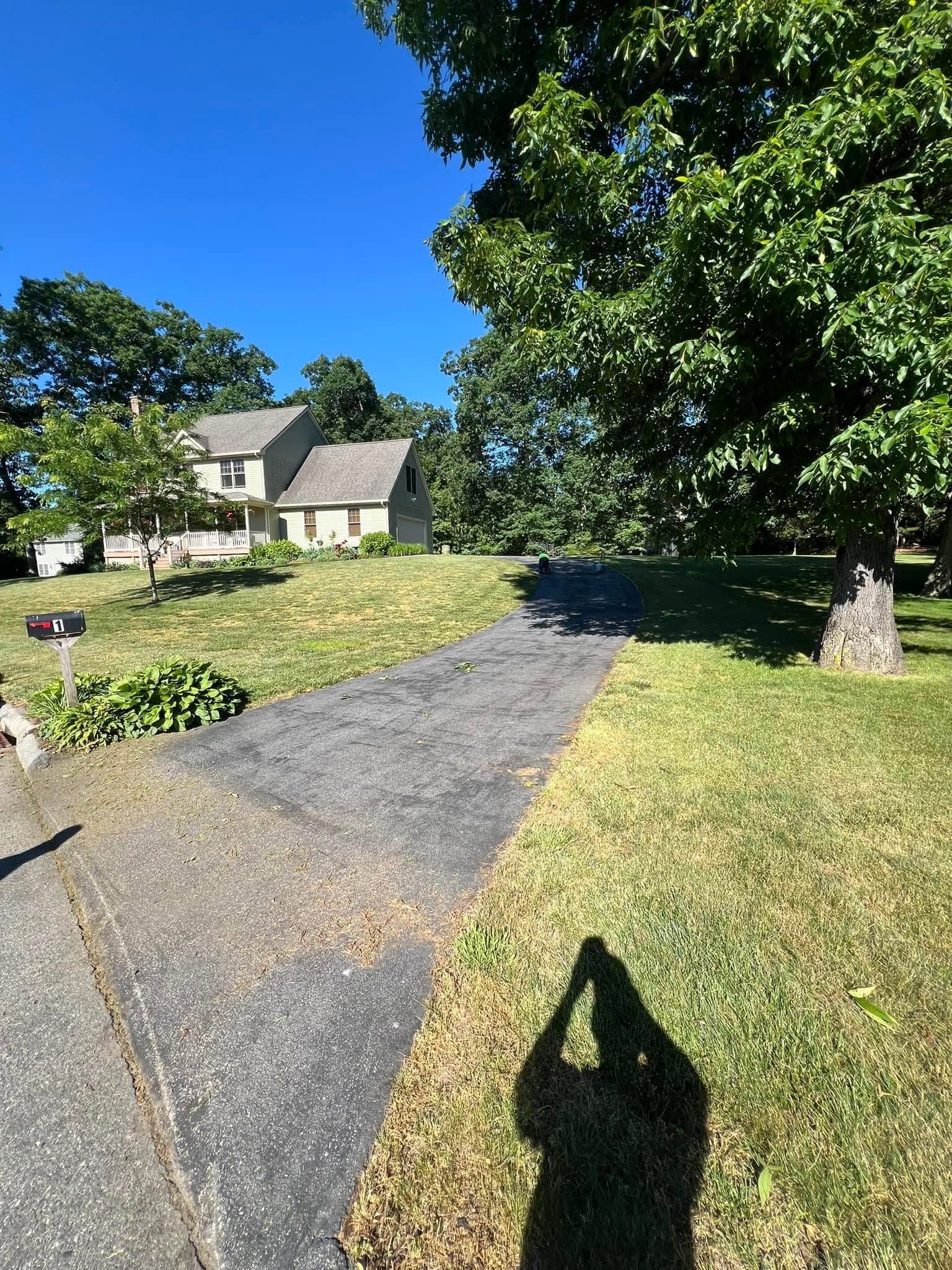 A paved driveway leads toward a two-story light-colored house surrounded by trees under a bright blue sky.