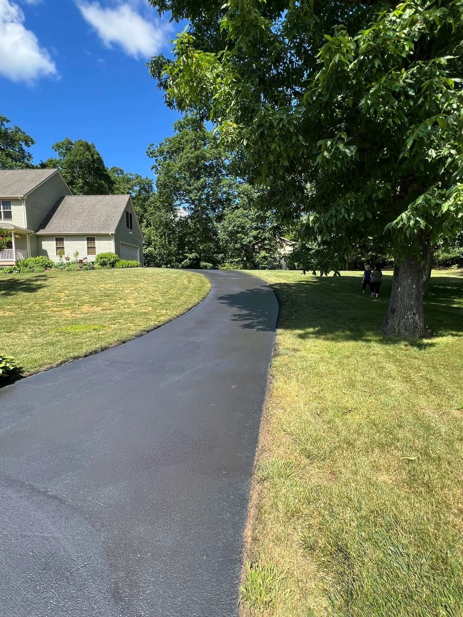 A dark asphalt driveway curves toward a suburban house surrounded by lush green trees under a bright blue sky.
