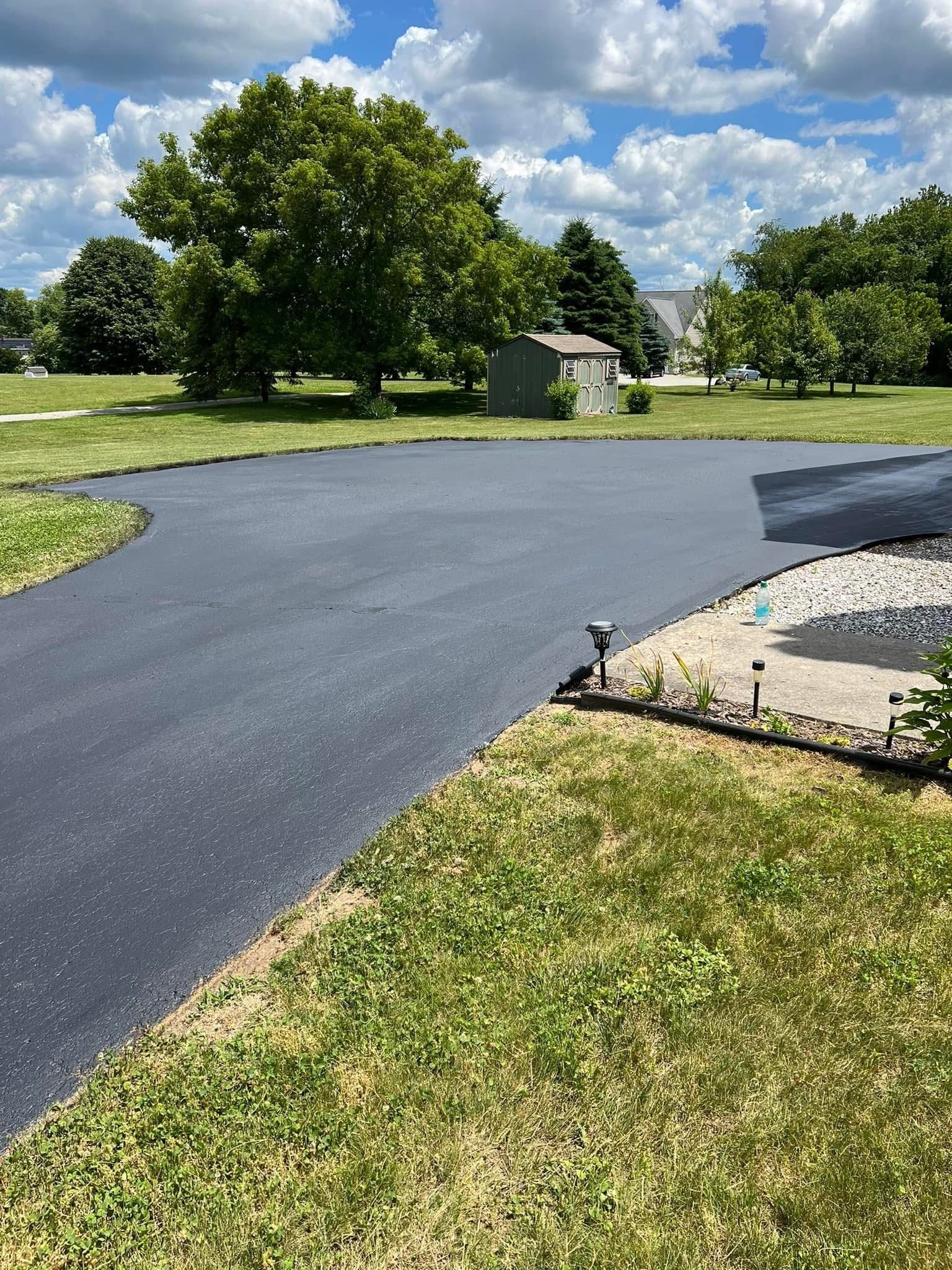 A freshly paved black asphalt driveway curves through a sunny green lawn with trees and a small shed in the background.