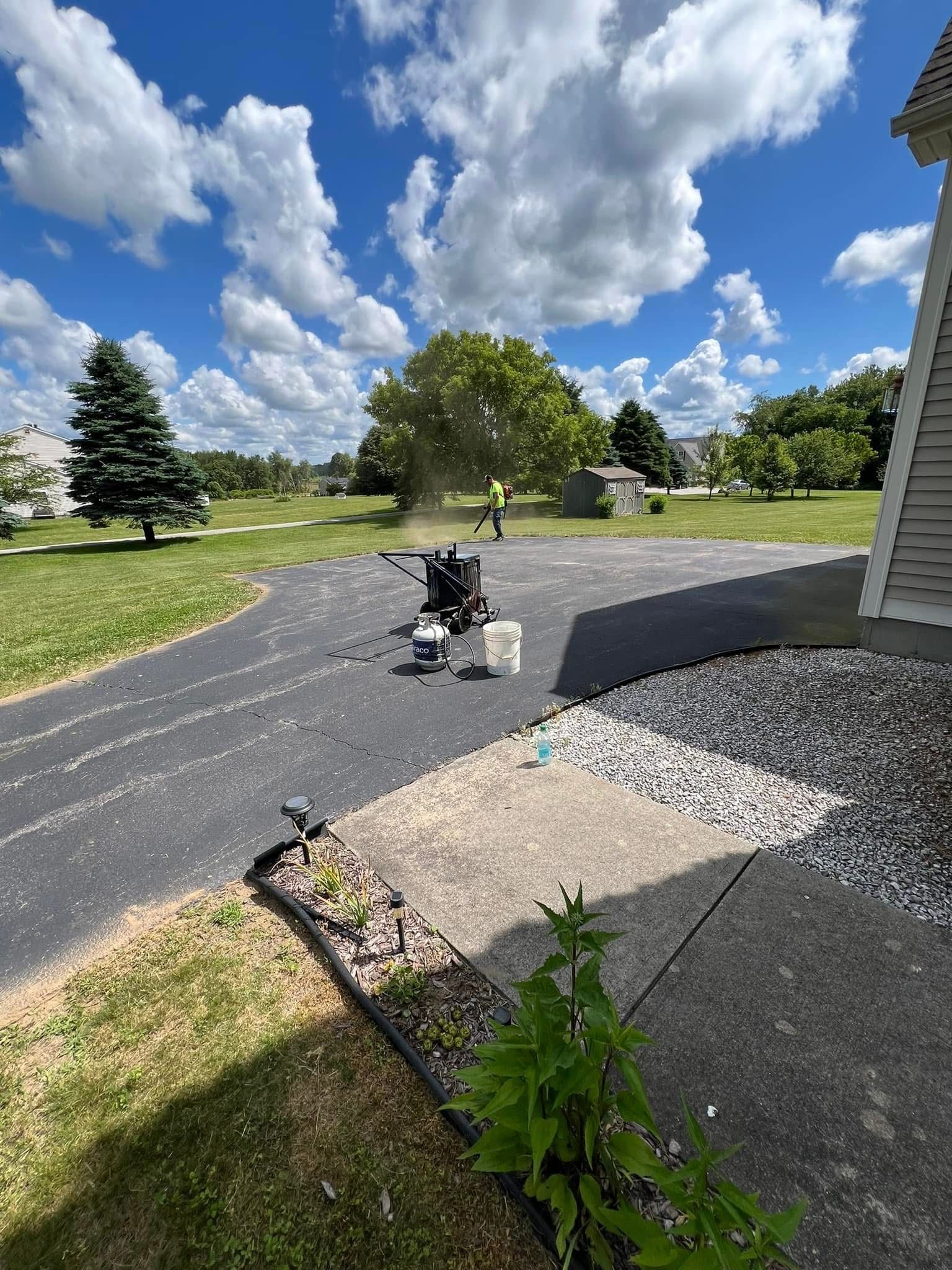A person uses a leaf blower to clear debris from a blacktop driveway on a sunny day with a blue, cloudy sky.
