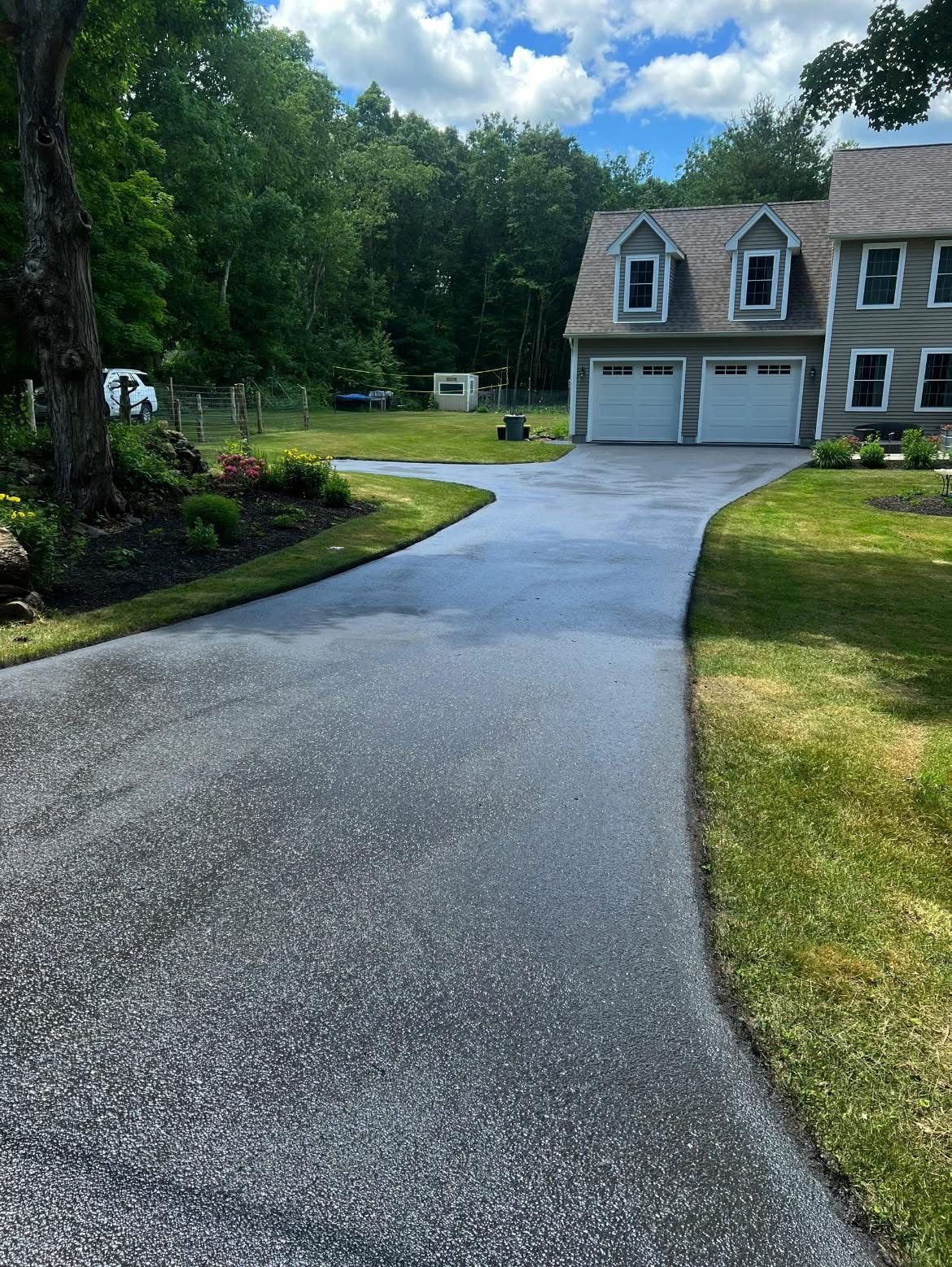 A gray two-story house with a two-car garage at the end of a long, paved driveway surrounded by trees and lawn.