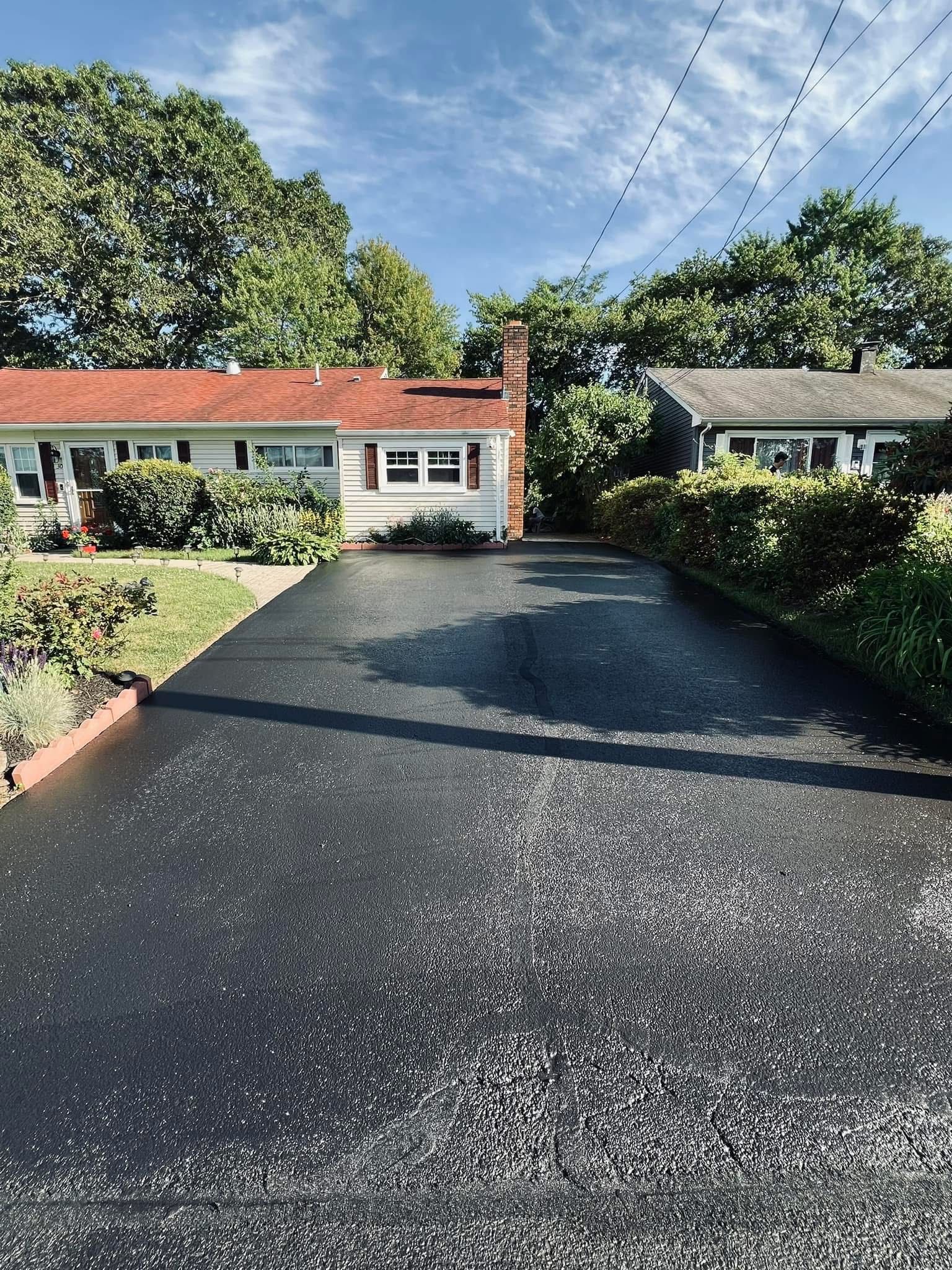 A freshly sealed black asphalt driveway leading toward a white house with a red roof under a bright blue sky.