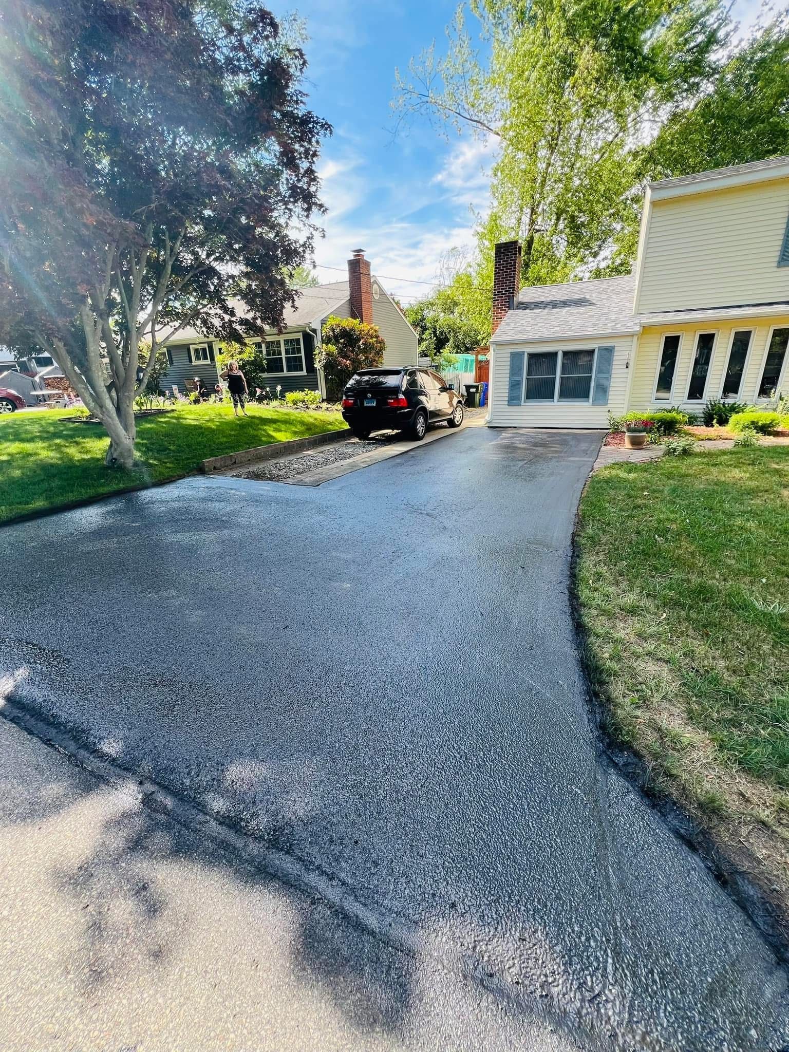 A freshly paved asphalt driveway leads up to a residential house on a sunny day with trees and a parked car nearby.