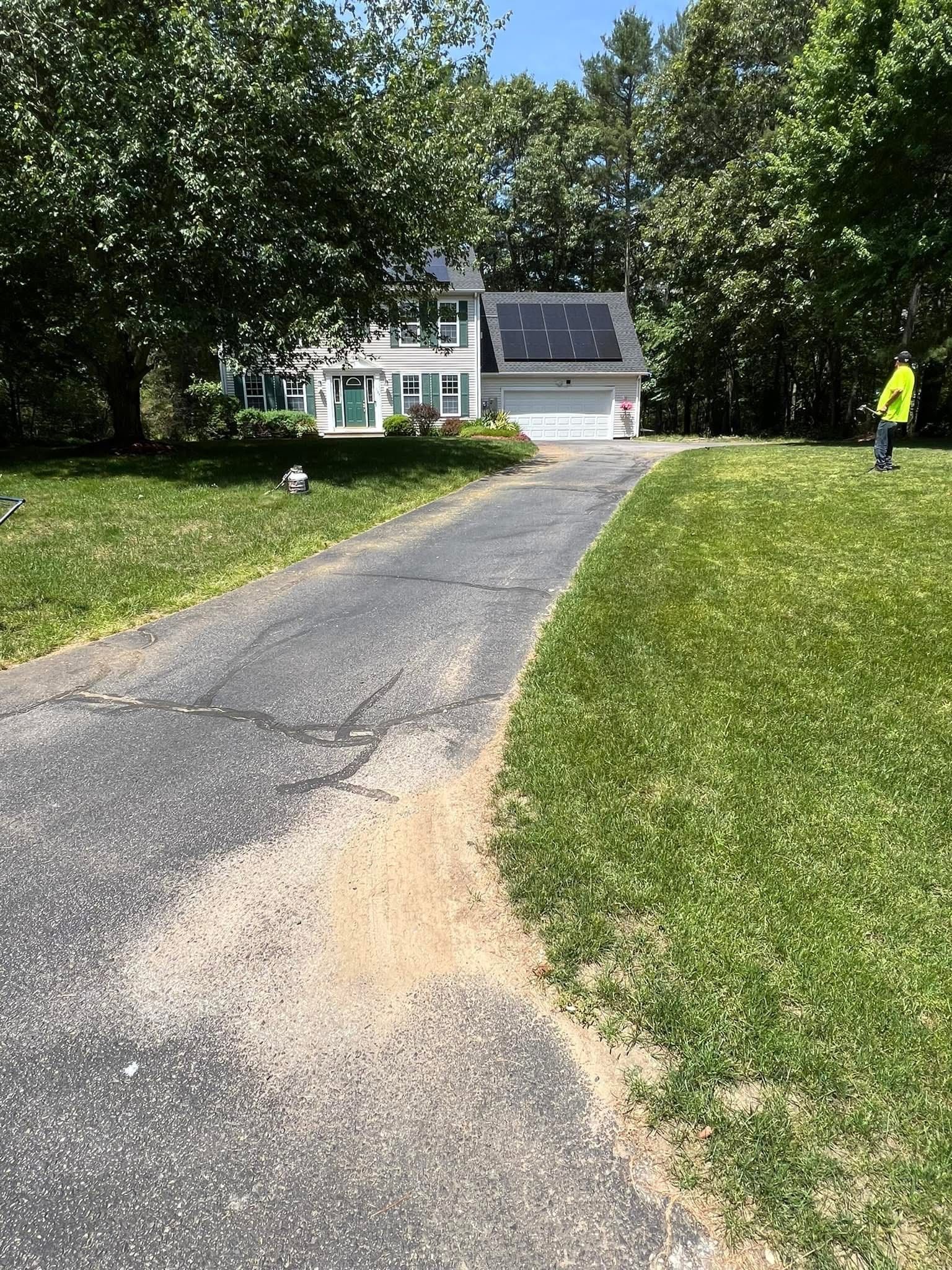 An asphalt driveway leads to a white house with solar panels on the roof, surrounded by trees and a green lawn.